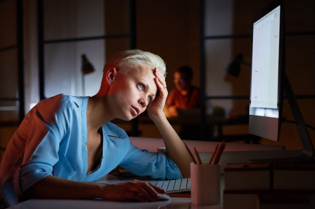 Woman sitting at a desk in front of a computer, resting her head on her hand and appearing tired or stressed while working late