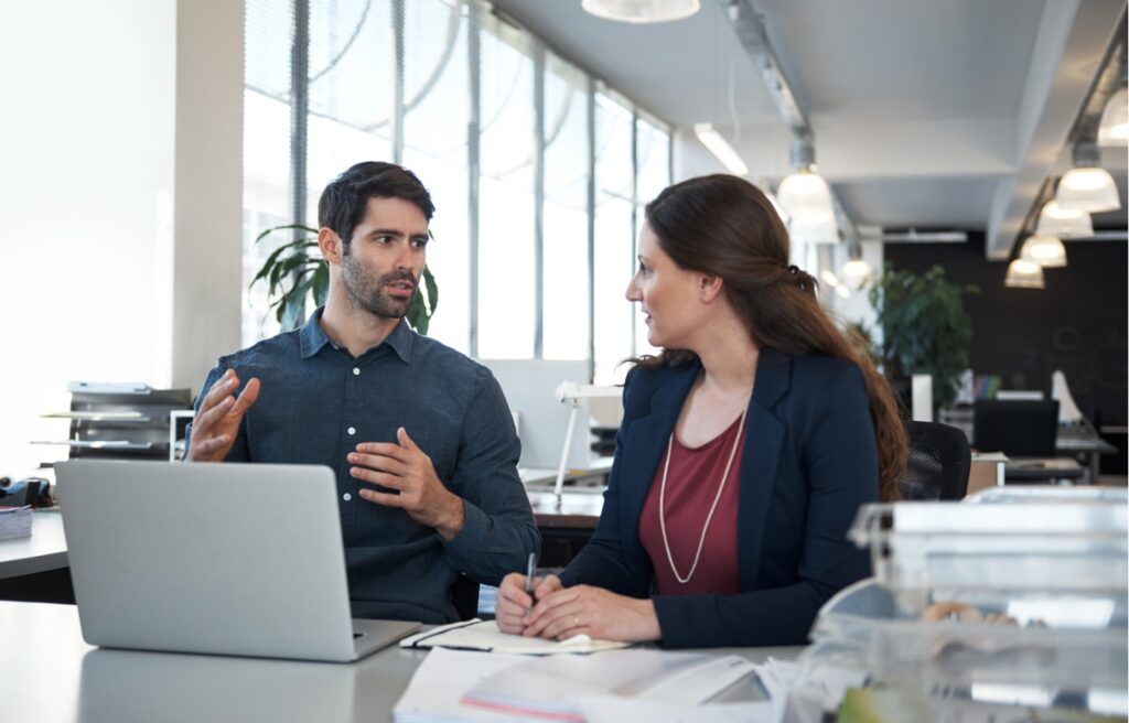 Two colleagues in a modern office discussing work at a desk, with one explaining a point while the other takes notes on a laptop
